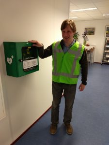 Kees, in BHV-unirorm, staat naast een AED.